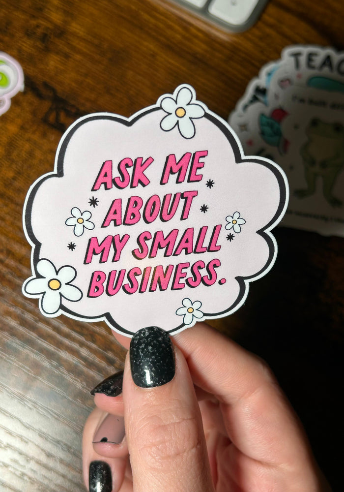 Sticker with 'Ask me about my small business' text held by a hand on a wooden surface.