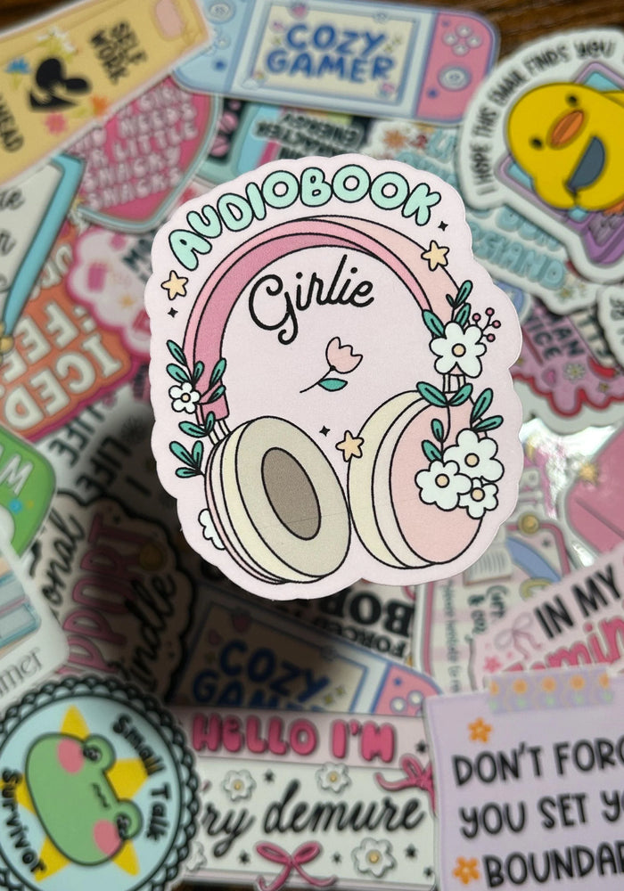 Mainly Pink Sticker with darker pink headphone, flowers and stars on the sides of the headphones and Text above the headphones saying Audiobook, Text inside the headphones saying Girlie. Sticker is in front of background of a lot of different stickers. 