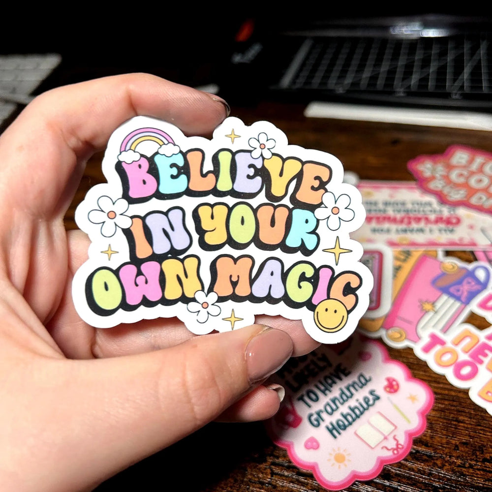 Hand holding a colourful sticker with 'Believe in your own magic' text on a wooden surface.