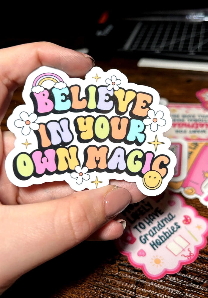 Hand holding a colourful sticker with 'Believe in your own magic' text on a wooden surface.