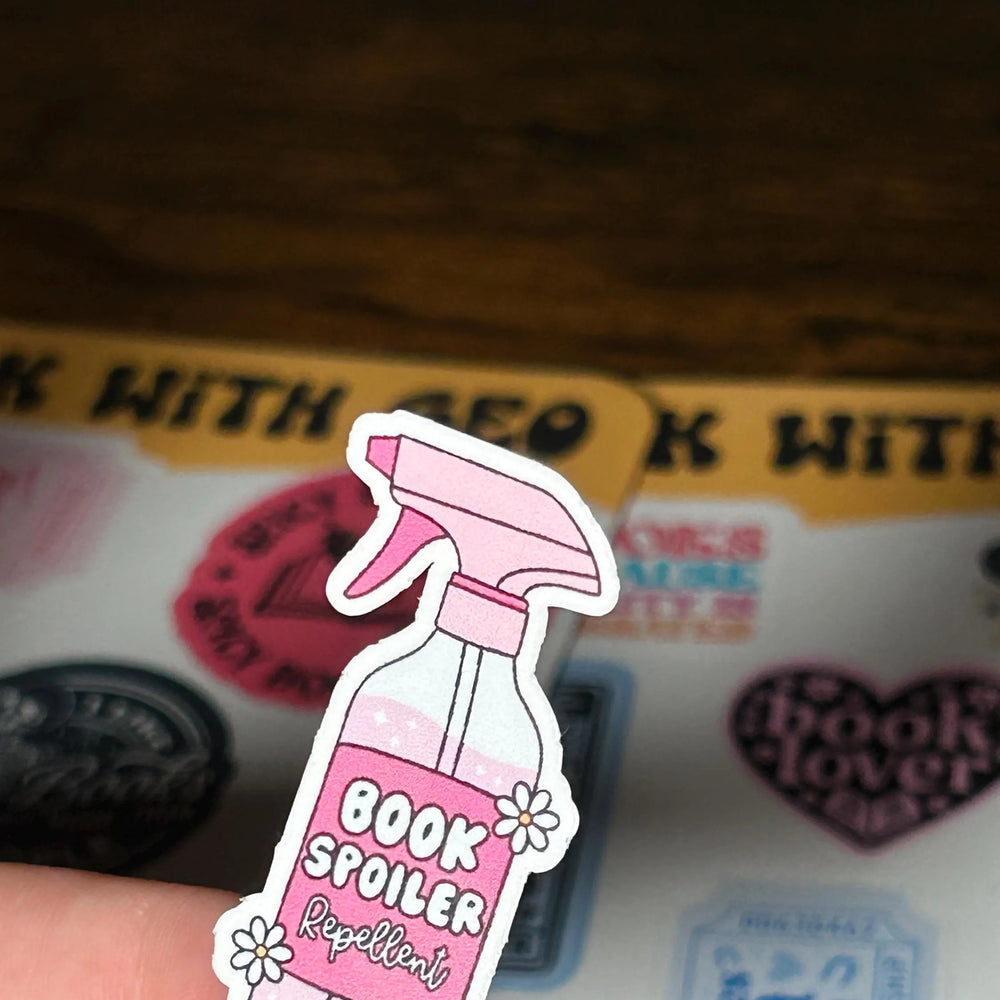 Pink 'Book Spoiler Repellent' sticker held over a book with stickers on a wooden surface.