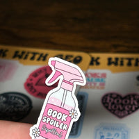 Load image into Gallery viewer, Pink 'Book Spoiler Repellent' sticker held over a book with stickers on a wooden surface.
