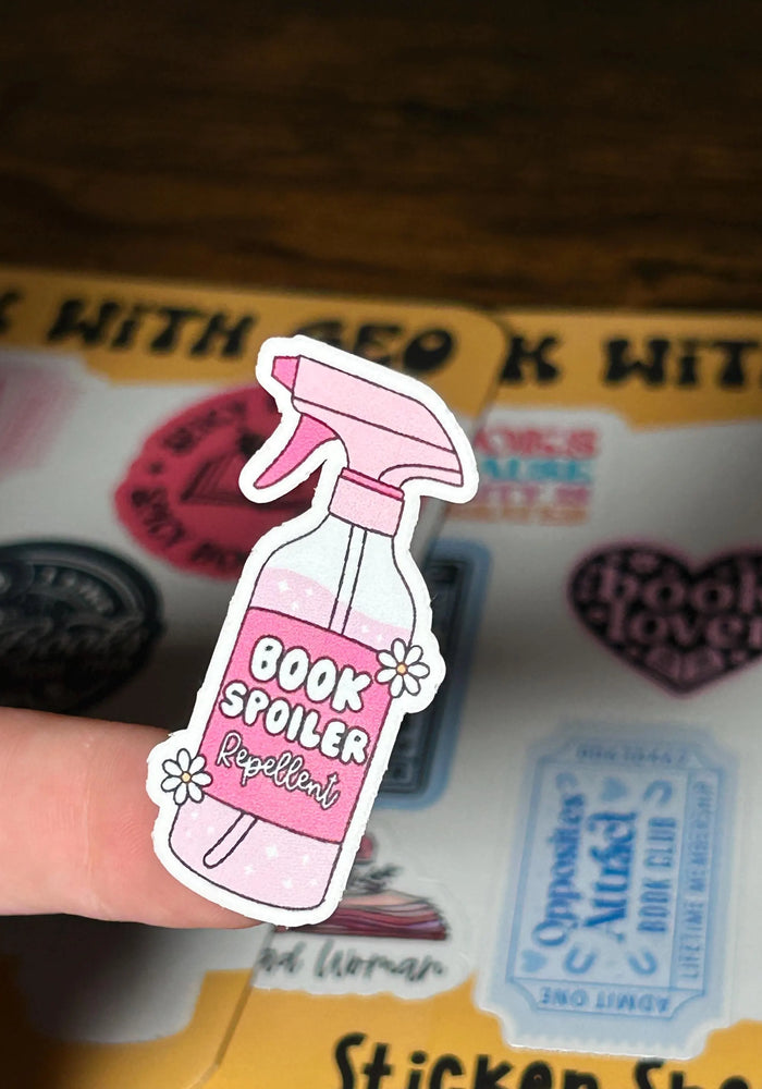 Pink 'Book Spoiler Repellent' sticker held over a book with stickers on a wooden surface.