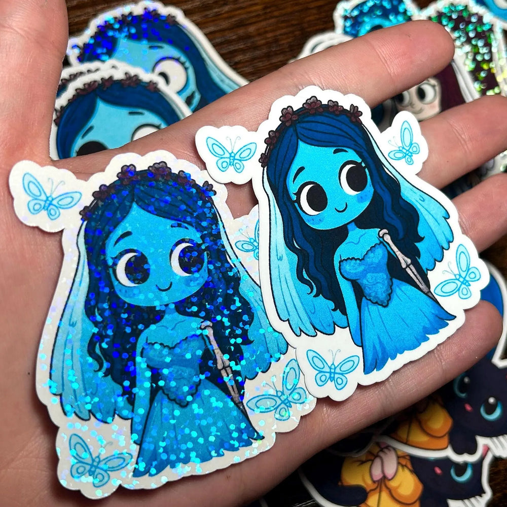 Hand holding two blue character stickers with butterfly designs, surrounded by more stickers on a wooden surface.