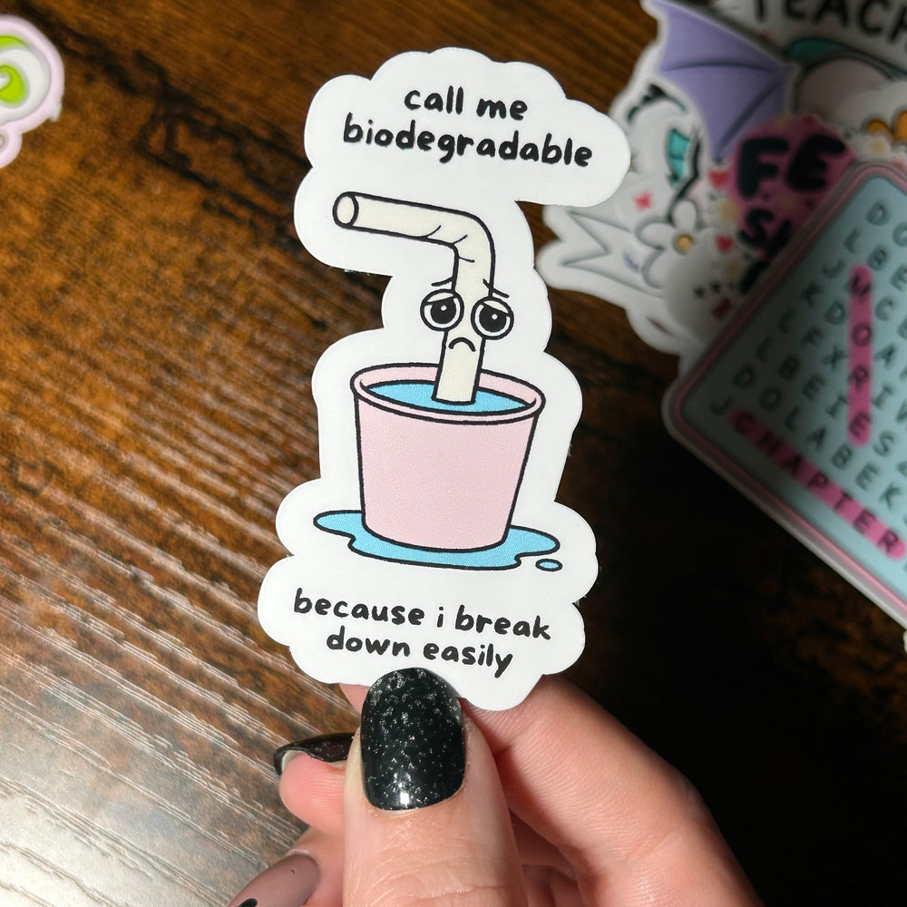 Sticker with a cartoon straw in a cup and humorous text, held by a hand on a wooden surface.