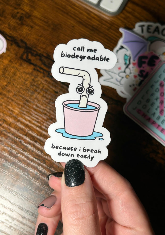 Sticker with a cartoon straw in a cup and humorous text, held by a hand on a wooden surface.