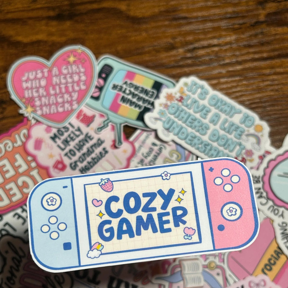 Sticker with 'Cozy Gamer' design on a wooden surface with other colourful stickers behind the Cozy Gamer Sticker.