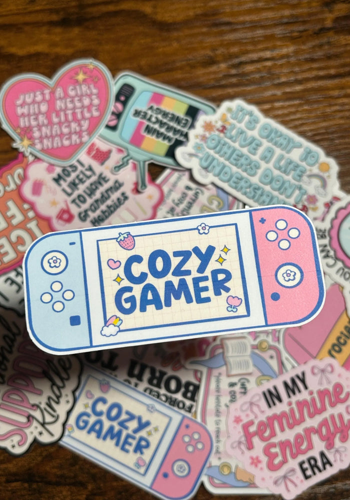 Sticker with 'Cozy Gamer' design on a wooden surface with other colourful stickers behind the Cozy Gamer Sticker.