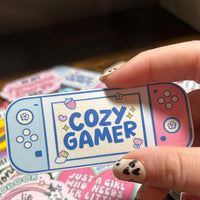 Load image into Gallery viewer, Hand holding a 'Cozy Gamer' product with other similar items in the background.
