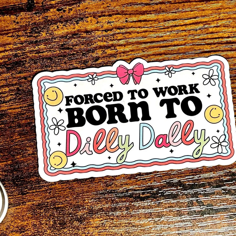 Sticker with text 'Forced to work, born to dilly dally' on a wooden surface