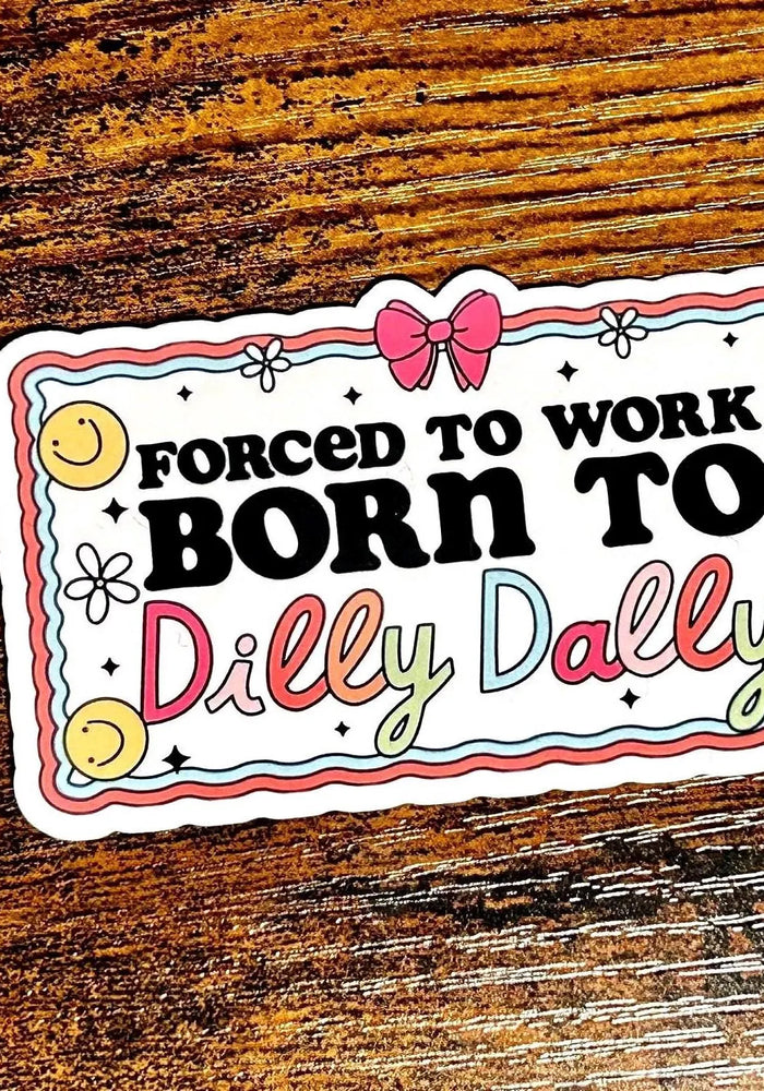 Sticker with text 'Forced to work, born to dilly dally' on a wooden surface