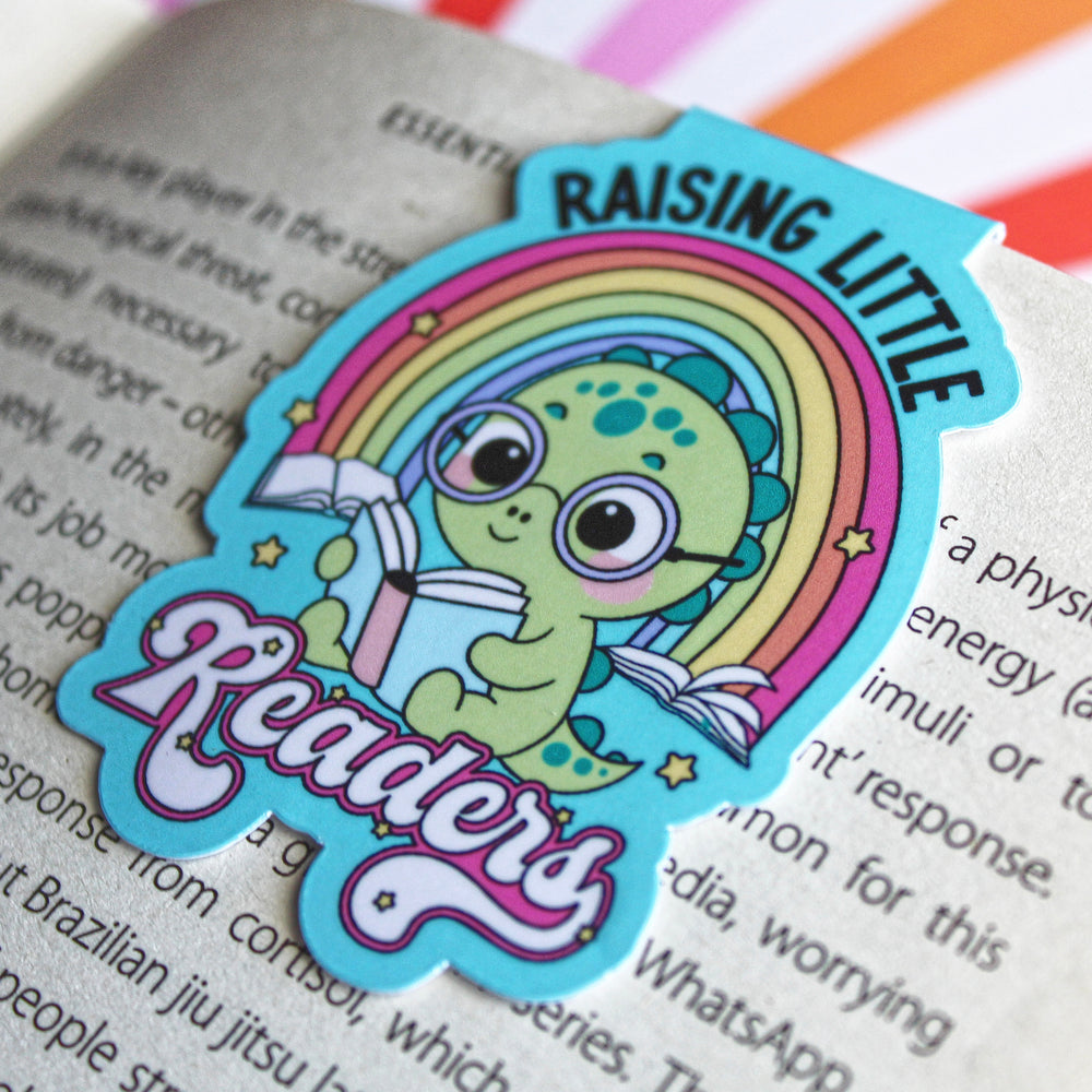 magnetic bookmark with a dinosaur holding books and a rainbow, with text 'Raising Little Readers', on a book page.