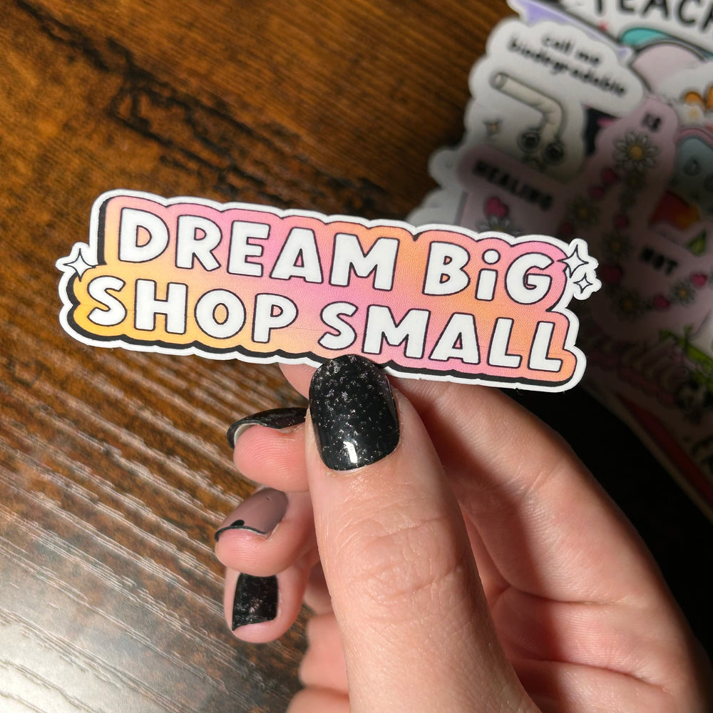 white text 'Dream big shop small' with gradient pink and yellow background on sticker, held by wooden table