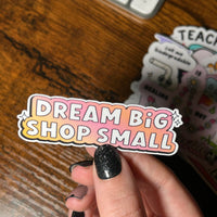 Load image into Gallery viewer, white text 'Dream big shop small' with gradient pink and yellow background on sticker, held by wooden table

