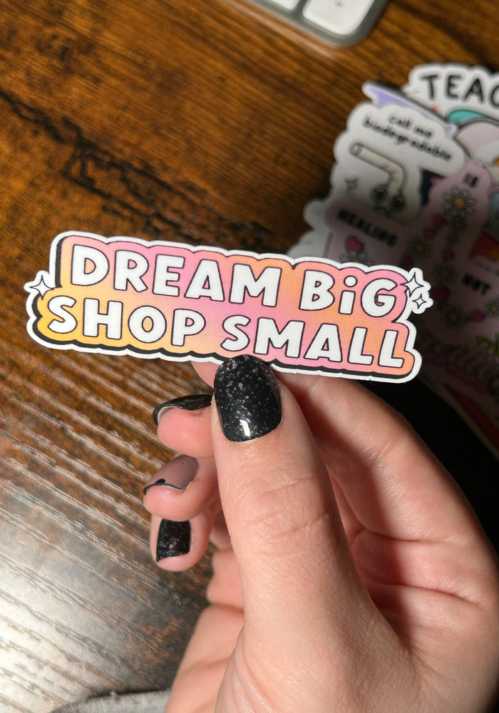 white text 'Dream big shop small' with gradient pink and yellow background on sticker, held by wooden table