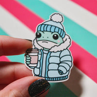 Load image into Gallery viewer, Cute Winter Frog Blue Coat Sticker – Cozy Frog in Puffer Jacket – Frog with Hot Drink – Glossy Waterproof Vinyl Sticker – Pastel Winter Aesthetic – 3.64cm x 5.5cm
