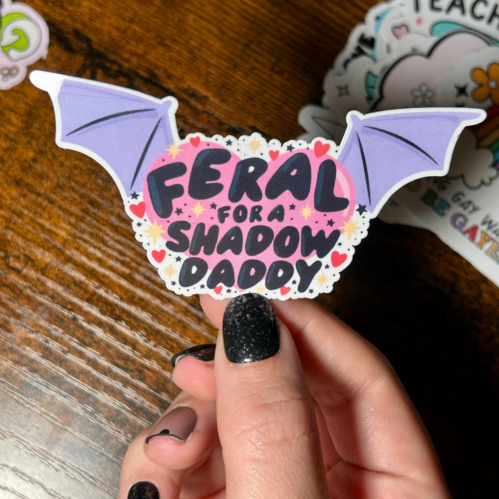 Sticker with text 'Feral for shadow daddy' with pink background and purple wings either side of the text, held in front of wooden table 