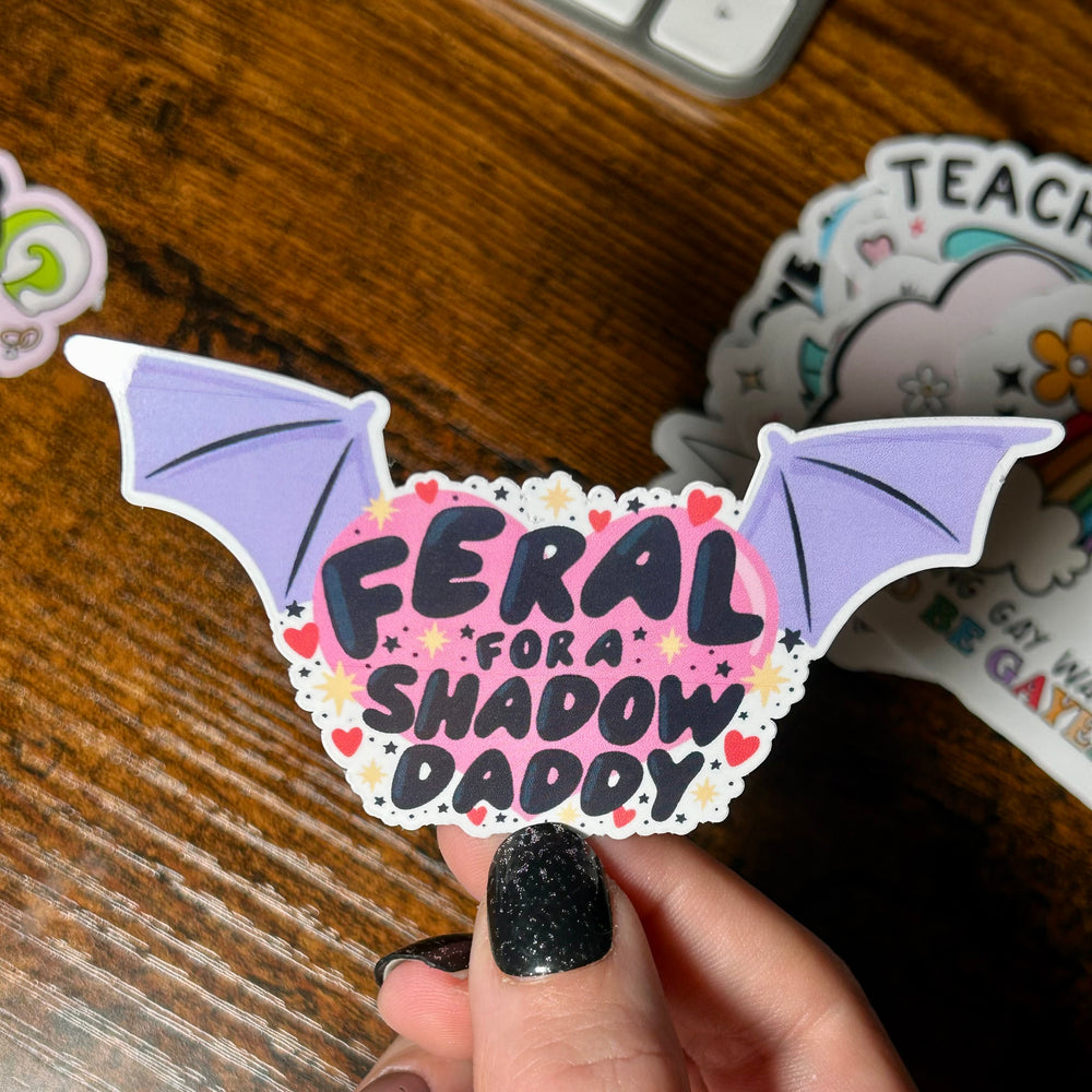 Sticker with text 'Feral for shadow daddy' with pink background and purple wings either side of the text, held in front of wooden table 