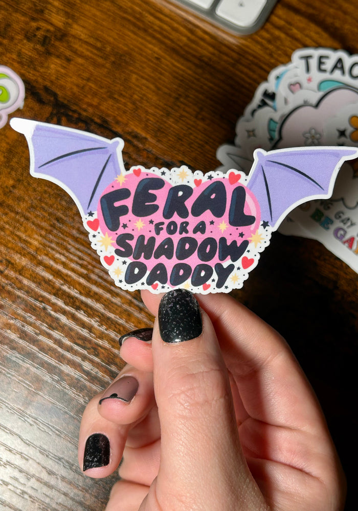 Sticker with text 'Feral for shadow daddy' with pink background and purple wings either side of the text, held in front of wooden table 