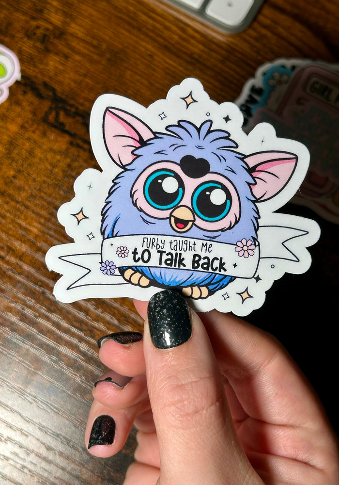 Sticker of a blue creature with text held by a hand on a wooden surface