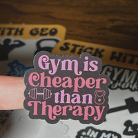 Load image into Gallery viewer, Sticker with 'Gym is cheaper than therapy' text held by a hand on a wooden surface.
