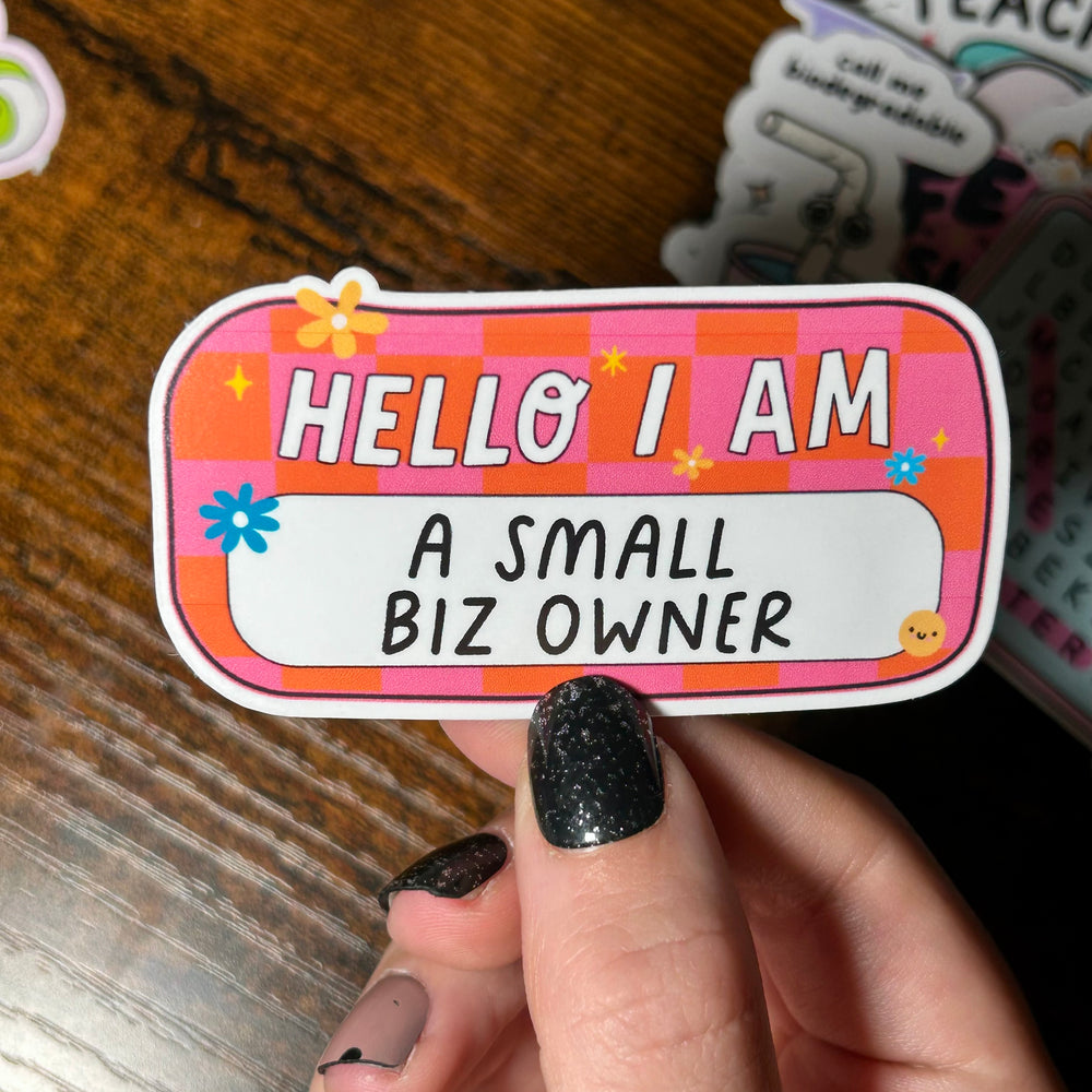 Hand holding a colorful sticker with text 'HELLO I AM A SMALL BIZ OWNER' on a wooden surface.