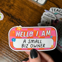 Load image into Gallery viewer, Hand holding a colorful sticker with text 'HELLO I AM A SMALL BIZ OWNER' on a wooden surface.
