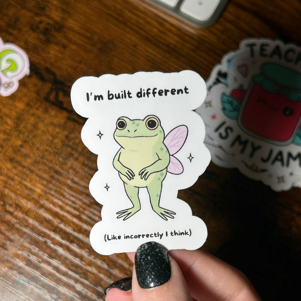 Sticker with a cartoon frog and text held by a hand on a wooden surface