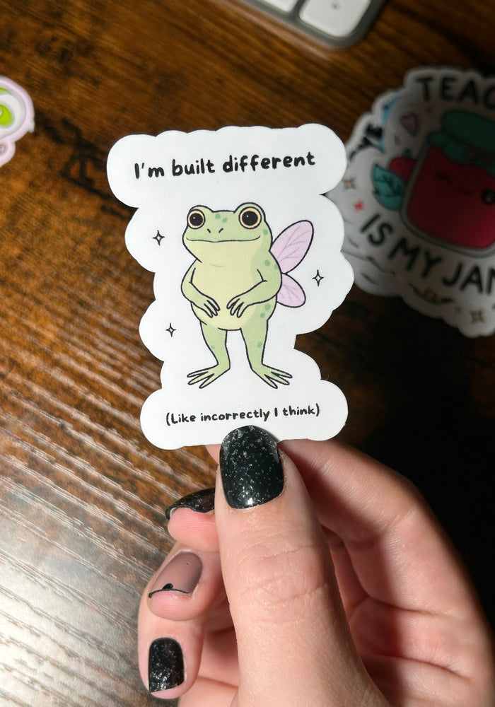 Sticker with a cartoon frog and text held by a hand on a wooden surface