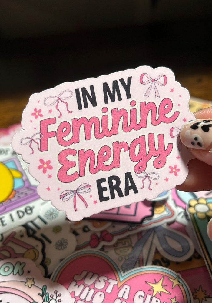 Sticker with 'In My Feminine Energy Era' text held by a hand on a colorful background