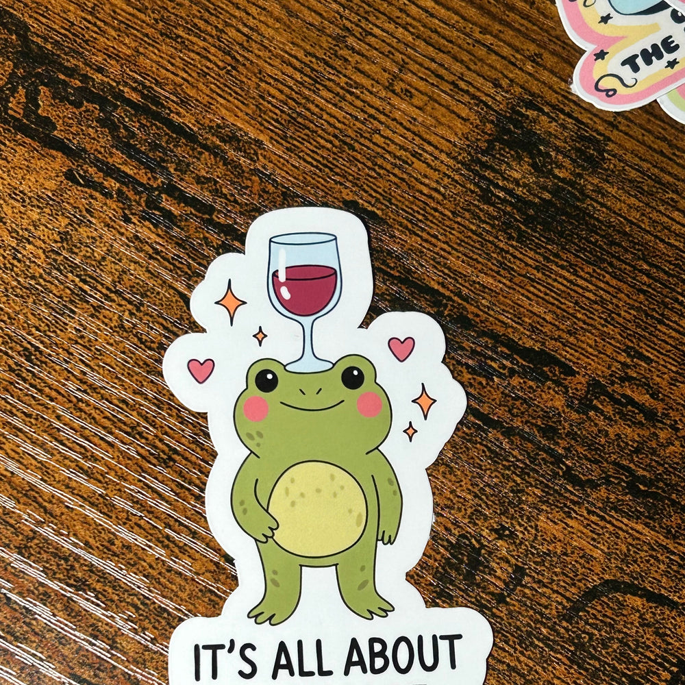 Sticker of a frog with a wine glass on its head and 'It's All About Balance' text on a wooden surface.