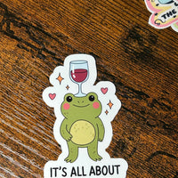 Load image into Gallery viewer, Sticker of a frog with a wine glass on its head and 'It's All About Balance' text on a wooden surface.
