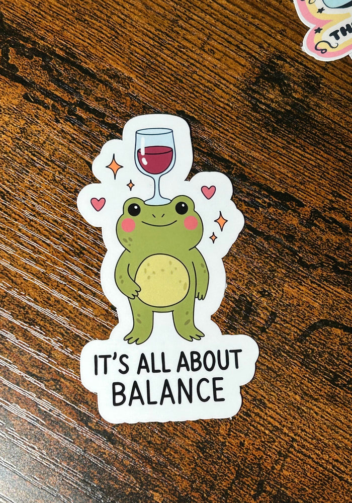 Sticker of a frog with a wine glass on its head and 'It's All About Balance' text on a wooden surface.