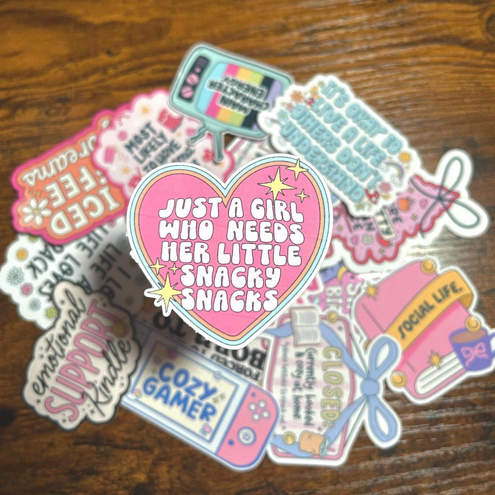Pink Sticker with text saying 'Just a girl who needs her little snacky snacks'. surrounded by collection of colourful stickers on a wooden surface