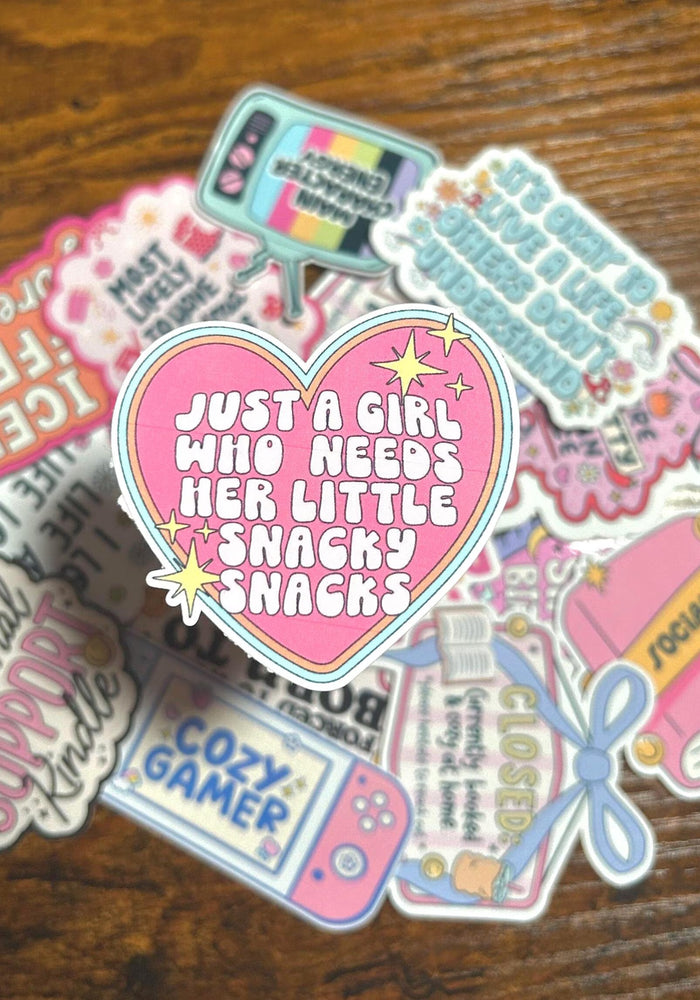 Pink Sticker with text saying 'Just a girl who needs her little snacky snacks'. surrounded by collection of colourful stickers on a wooden surface
