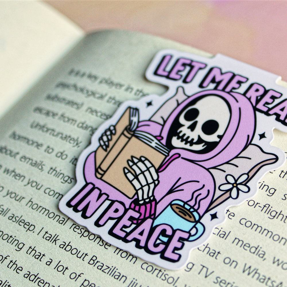 bookmark with a skeleton holding books and coffee, text 'Let me read in peace', on an open book.