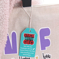 Load image into Gallery viewer, Beige tote bag with purple cut-out letters and a green tag reading 'Stick with Geo'.
