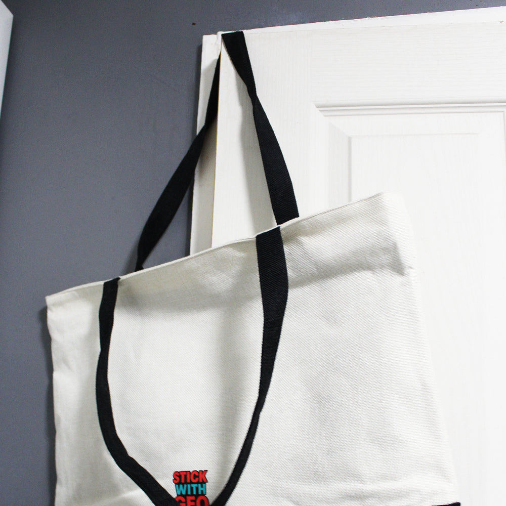 White tote bag with black handles and trim, featuring a logo, on a white surface.