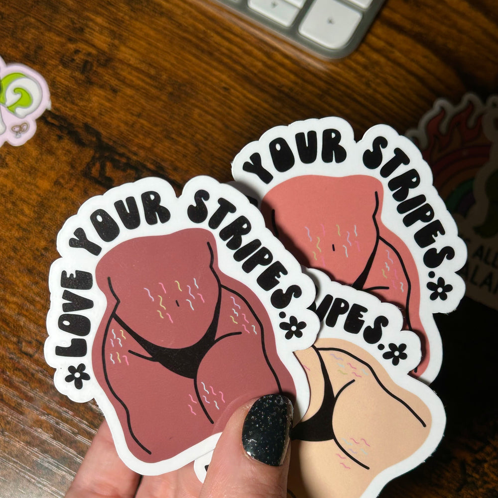 Two stickers with cartoon illustrations and text held by a hand on a wooden surface.