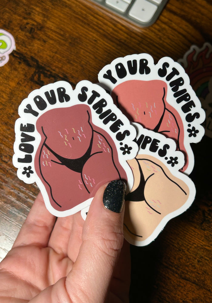Two stickers with cartoon illustrations and text held by a hand on a wooden surface.