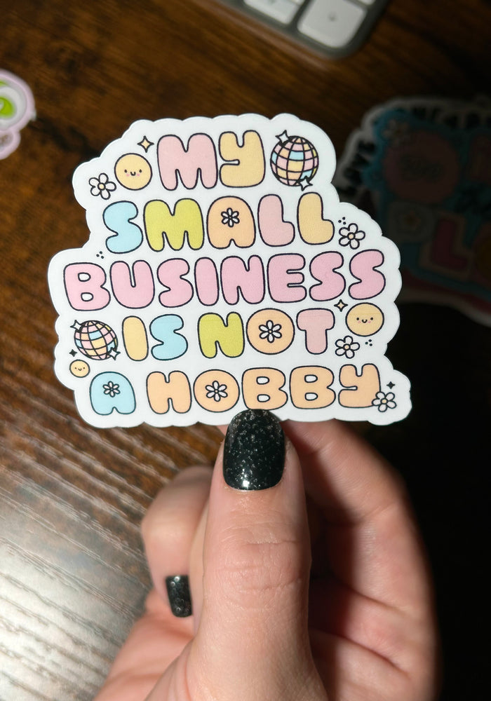 Sticker with 'My small business is not a hobby' text held by a hand on a wooden surface.