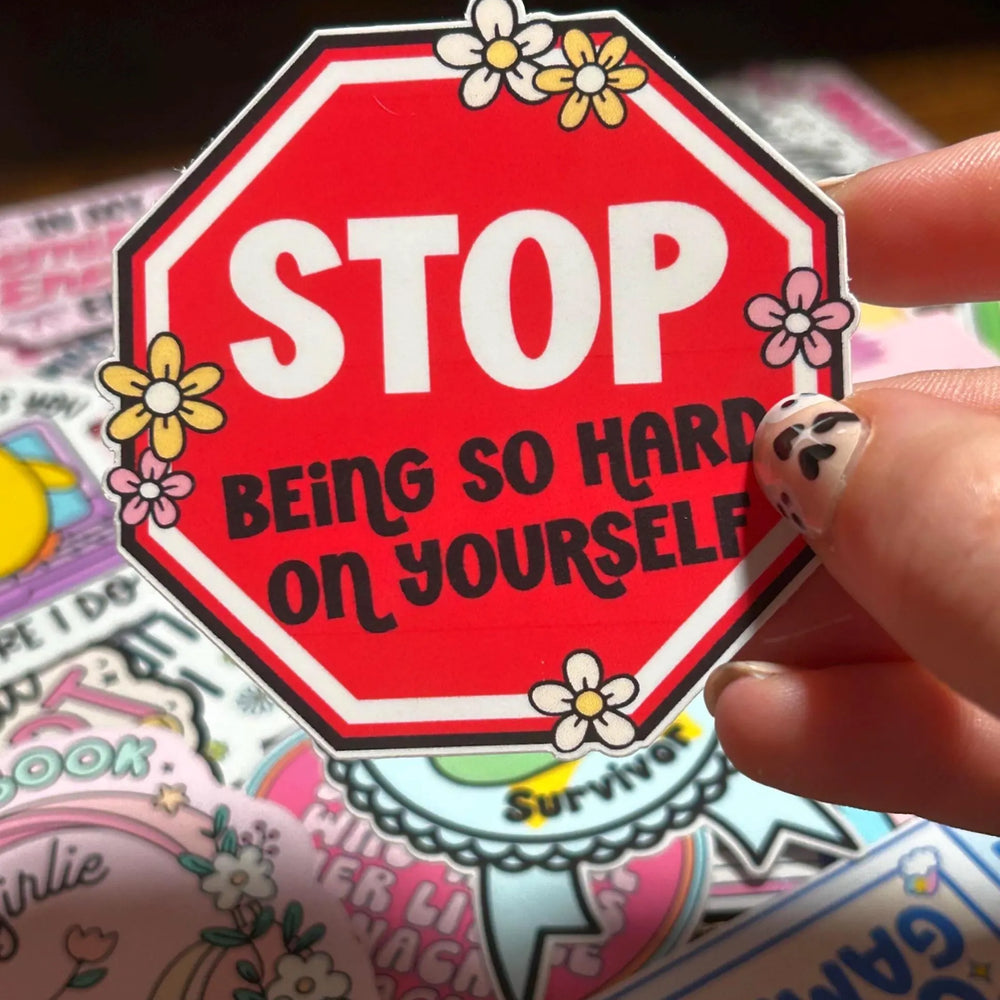 Sticker with 'STOP BEING SO HARD ON YOURSELF' text held by a hand