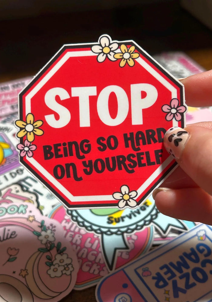 Sticker with 'STOP BEING SO HARD ON YOURSELF' text held by a hand