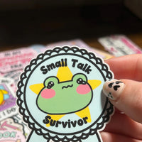 Load image into Gallery viewer, Sticker with a frog design and 'Small Talk Survivor' text held by a hand.
