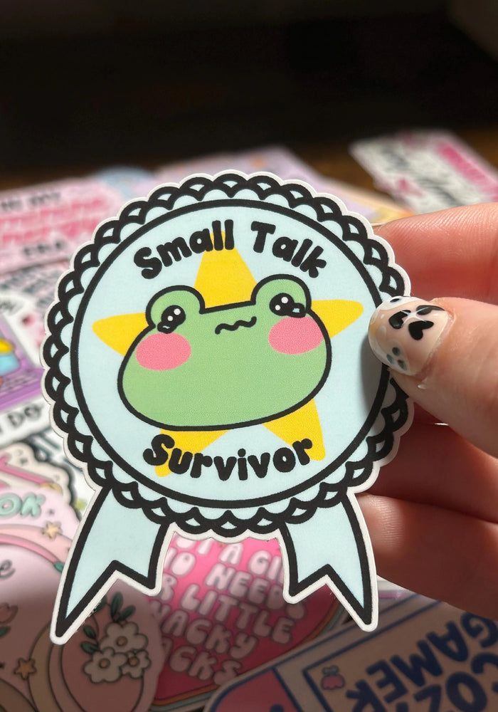 Sticker with a frog design and 'Small Talk Survivor' text held by a hand.