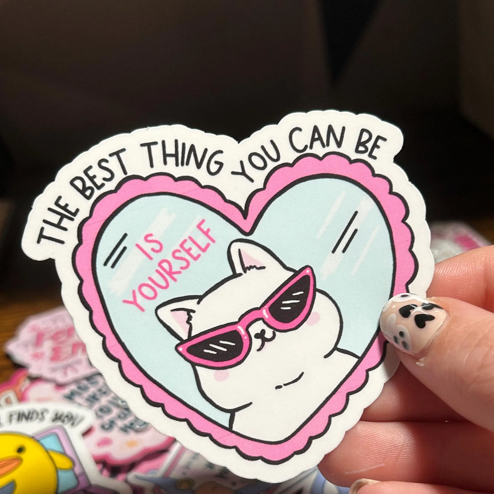 Sticker with a cat wearing sunglasses and text 'The best thing you can be is yourself' held by a hand.