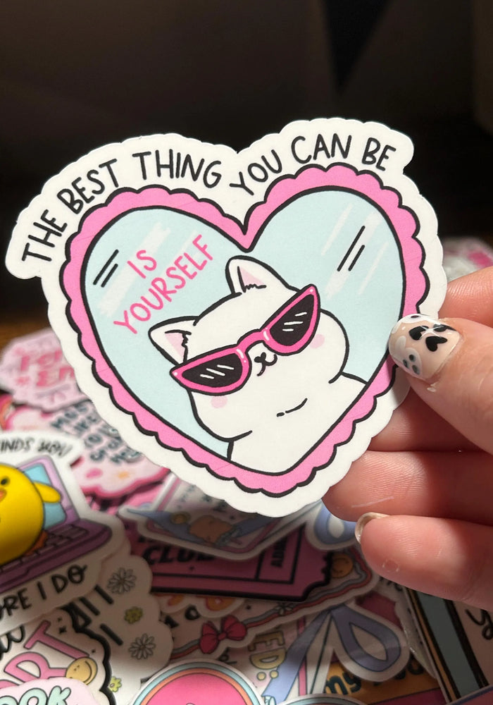Sticker with a cat wearing sunglasses and text 'The best thing you can be is yourself' held by a hand.