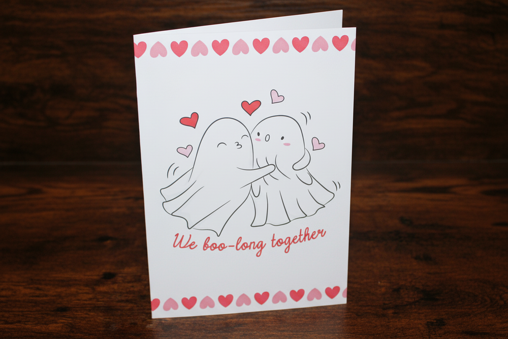 Greeting card with ghost couple illustration and 'We boo-long together' text on a wooden surface