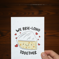 Load image into Gallery viewer, Card with a brie illustration and text 'We brie-long together' held by a hand on a wooden surface

