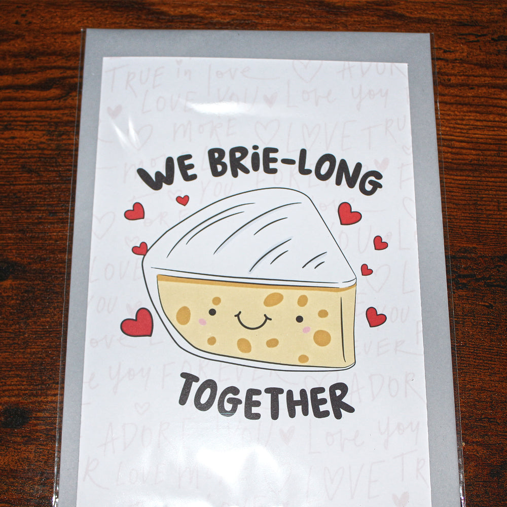 Card with a cartoon brie cheese and text 'We brie-long together' on a wooden surface